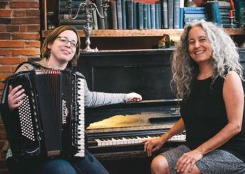 Two people sit at near a piano with one holding an accordion.